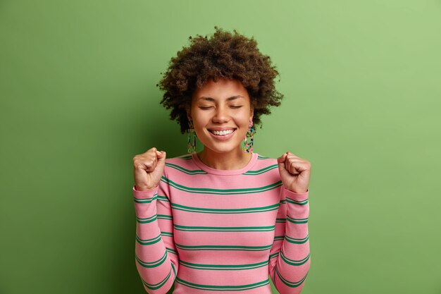 Feliz joven aprieta los puños celebra el éxito cierra los ojos y sonríe ampliamente espera el anuncio de los resultados vestida con un suéter a rayas aislado sobre una pared verde vivo