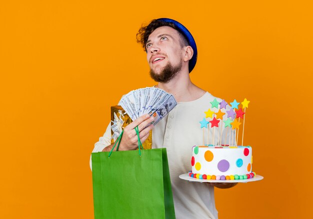 Feliz joven apuesto chico de fiesta eslavo con sombrero de fiesta con caja de regalo bolsa de papel de dinero y pastel de cumpleaños con estrellas mirando hacia arriba soñando aislado sobre fondo naranja con espacio de copia