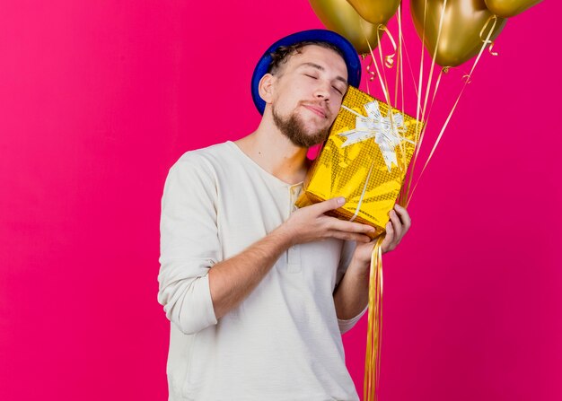 Feliz joven apuesto chico de fiesta eslavo con sombrero de fiesta sosteniendo globos y tocando la cara con caja de regalo con los ojos cerrados aislado sobre fondo carmesí con espacio de copia