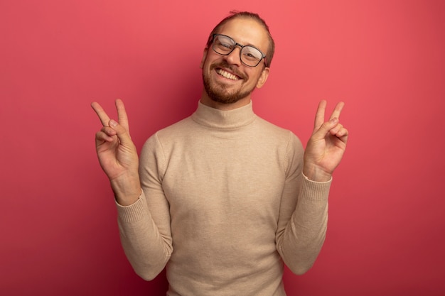 Feliz joven apuesto hombre de cuello alto beige y gafas mirando al frente sonriendo alegremente mostrando el signo v con ambas manos de pie sobre la pared rosa