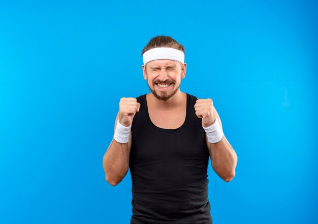 Feliz joven apuesto hombre deportivo con diadema y muñequeras apretando los puños con los ojos cerrados aislados en la pared azul con espacio de copia