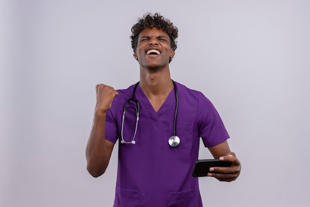 Feliz joven apuesto hombre de piel oscura con cabello rizado en uniforme médico violeta sosteniendo su teléfono inteligente con el puño apretado