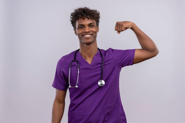 Un feliz joven apuesto médico de piel oscura con cabello rizado vistiendo uniforme violeta con estetoscopio mostrando gesto de fuerza