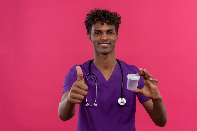Un feliz joven apuesto médico de piel oscura con cabello rizado vistiendo uniforme violeta con estetoscopio mostrando los pulgares hacia arriba mientras sostiene un frasco de plástico para muestras