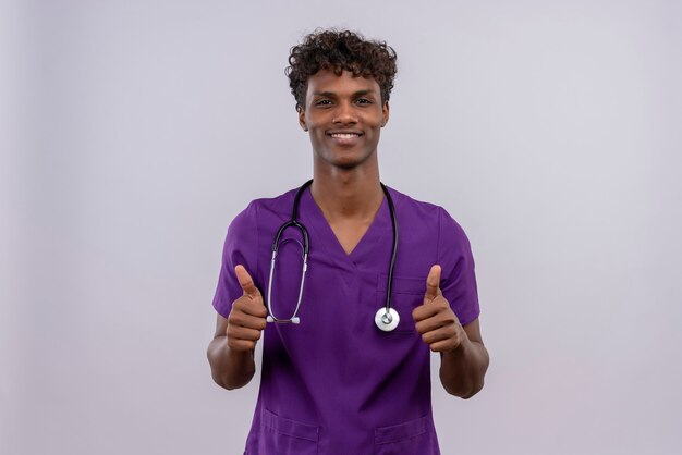 Un feliz joven apuesto médico de piel oscura con cabello rizado vistiendo uniforme violeta con estetoscopio mostrando los pulgares para arriba