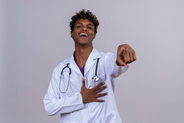 Un feliz joven apuesto médico de piel oscura con pelo rizado vistiendo bata blanca con estetoscopio apuntando a la cámara con los dedos índices