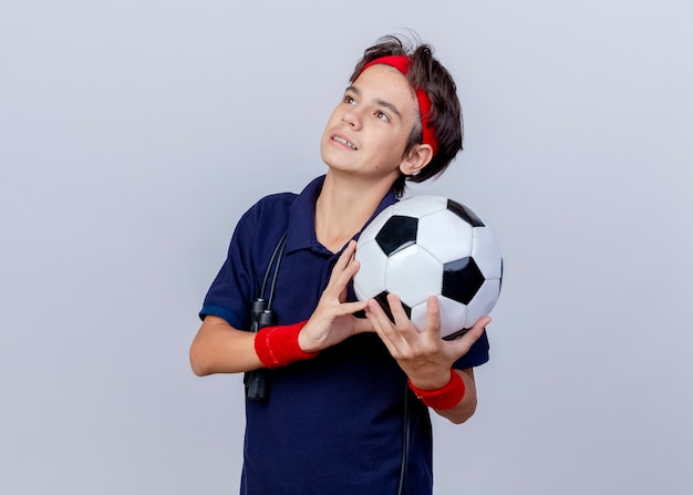 Feliz joven apuesto muchacho deportivo con diadema y muñequeras con aparatos dentales y saltar la cuerda alrededor del cuello sosteniendo un balón de fútbol mirando hacia arriba aislado sobre fondo blanco con espacio de copia