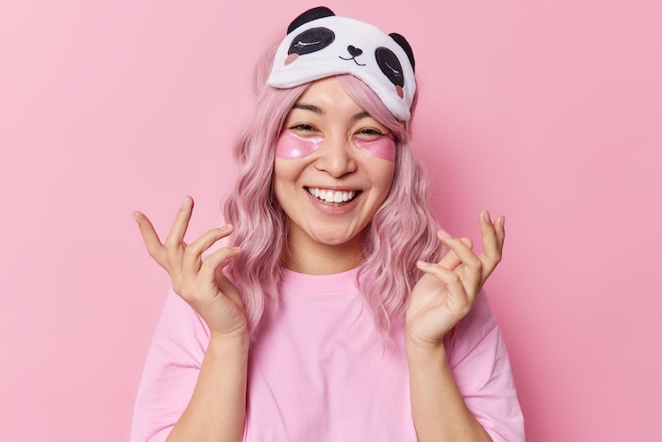 Foto gratuito feliz joven asiática con el pelo teñido de rosa sonríe con dientes aplica parches de hidrogel debajo de los ojos mantiene las manos levantadas disfruta de los procedimientos de belleza después de dormir con los ojos vendados en la frente y la camiseta