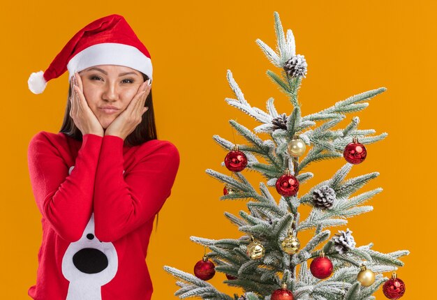 Feliz joven asiática vistiendo gorro de navidad con suéter de pie cerca del árbol de navidad poniendo las manos en las mejillas aisladas sobre fondo naranja