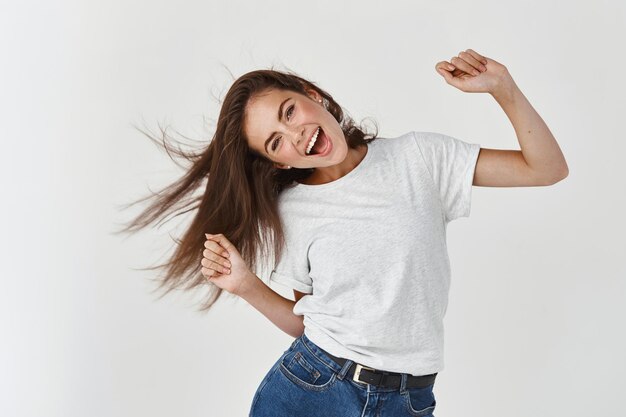 Feliz joven bailando y látigo cabello sano y brillante, divirtiéndose, de pie alegre sobre la pared blanca