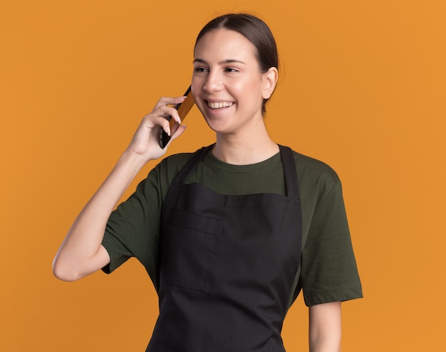 Feliz joven barbero morena en uniforme habla por teléfono mirando al lado en naranja