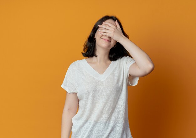 Feliz joven bastante caucásica con gafas cerrando los ojos con la mano aislada sobre fondo naranja con espacio de copia