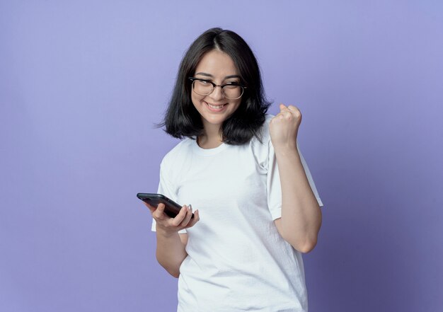 Feliz joven bastante caucásica con gafas mirando al lado sosteniendo el teléfono móvil y apretando el puño aislado sobre fondo púrpura con espacio de copia