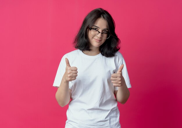 Feliz joven bastante caucásica con gafas mostrando los pulgares hacia arriba en la cámara aislada sobre fondo carmesí con espacio de copia