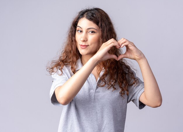 Feliz joven bastante caucásica haciendo signo de corazón aislado en la pared blanca