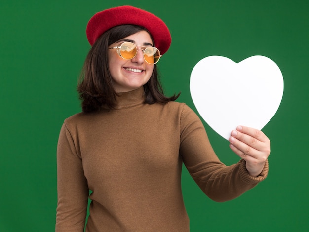 Foto gratuita feliz joven bastante caucásica con sombrero de boina en gafas de sol con forma de corazón en verde