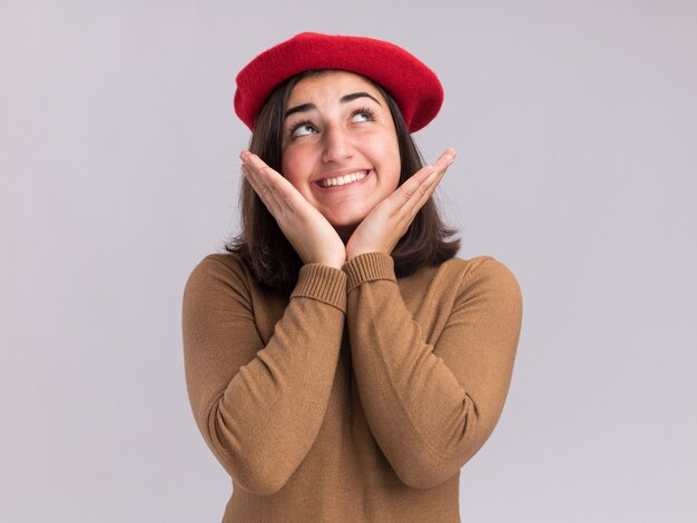 Feliz joven bastante caucásica con sombrero de boina pone las manos en la barbilla y mira hacia arriba