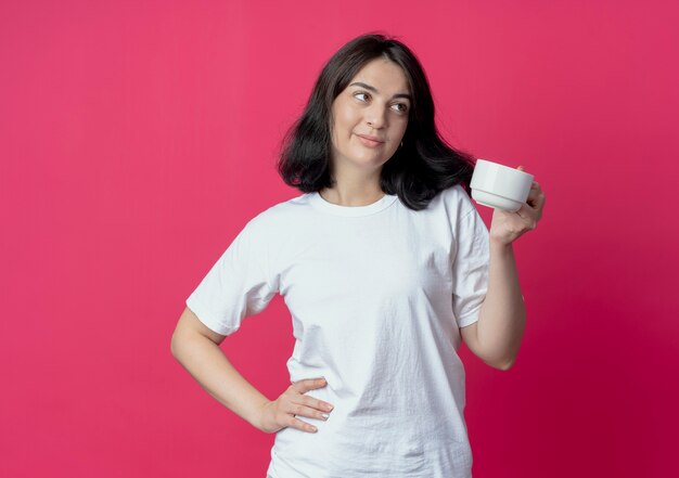 Feliz joven bastante caucásica sosteniendo la taza manteniendo la mano en la cintura y mirando al lado aislado sobre fondo carmesí con espacio de copia