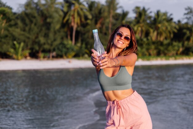 Feliz joven y bella mujer europea con una botella de agua de plástico en la mano en la playa