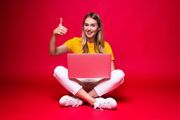 Feliz joven bella mujer rizada sentada en el suelo con las piernas cruzadas y usando la computadora portátil en la pared roja.