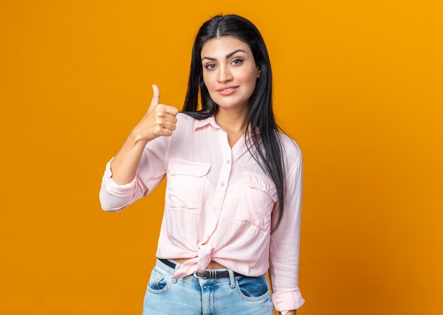 Feliz joven y bella mujer en ropa casual mirando al frente sonriendo confiado mostrando los pulgares para arriba de pie sobre la pared naranja