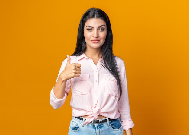 Feliz joven y bella mujer en ropa casual sonriendo confiado mostrando los pulgares para arriba de pie sobre la pared naranja