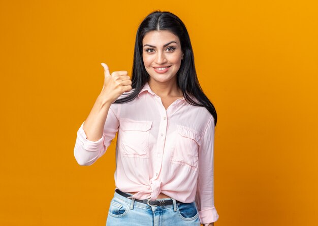 Feliz joven y bella mujer en ropa casual sonriendo confiado mostrando los pulgares para arriba de pie sobre la pared naranja