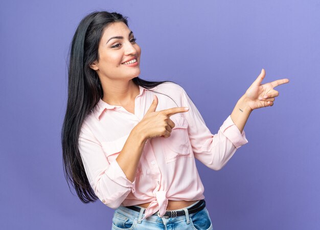 Feliz joven y bella mujer en ropa casual sorprendida señalando con los dedos índices hacia el lado de pie sobre la pared azul