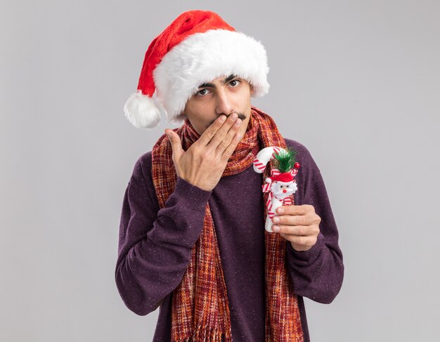 Feliz joven bigotudo vistiendo gorro de Papá Noel de Navidad con bufanda alrededor de su cuello sosteniendo bastón de caramelo de Navidad soplando un beso