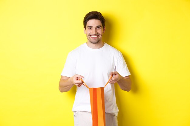 Feliz joven bolsa de compras abierta con presente, sonriendo a la cámara, de pie contra el fondo amarillo.