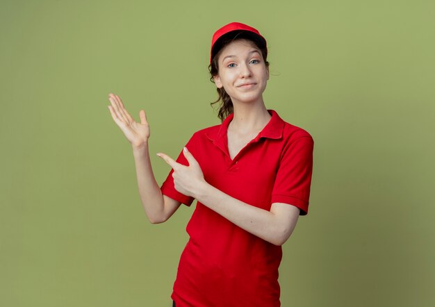 Feliz joven bonita repartidora en uniforme rojo y gorra mostrando la mano vacía y apuntando al lado aislado sobre fondo verde oliva con espacio de copia