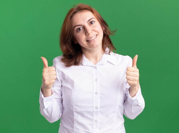 Feliz joven con camisa blanca mirando al frente con una sonrisa en la cara mostrando los pulgares para arriba de pie sobre la pared verde