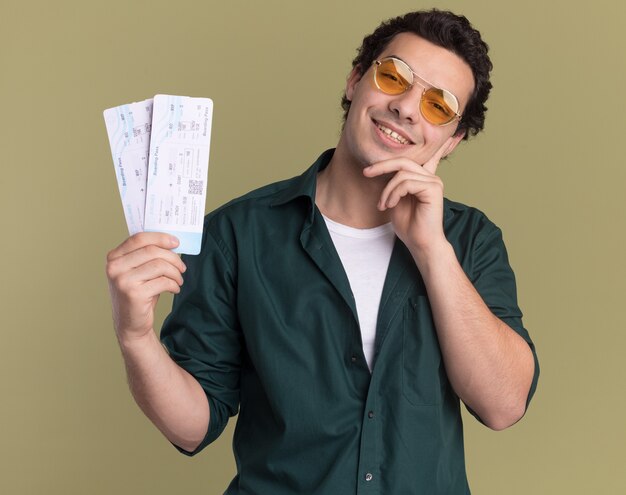 Feliz joven en camisa verde con gafas con billetes de avión mirando al frente con una sonrisa en la cara de pie sobre la pared verde