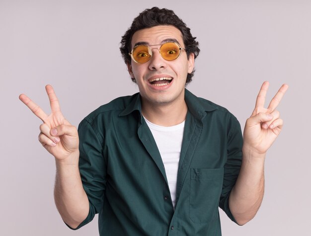 Feliz joven en camisa verde con gafas mirando al frente sonriendo alegremente mostrando v-sign de pie sobre la pared blanca