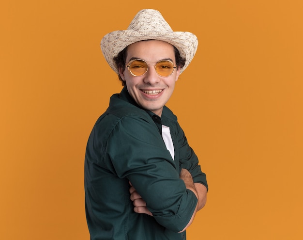 Foto gratuita feliz joven en camisa verde y sombrero de verano con gafas mirando al frente con una sonrisa en la cara de pie sobre la pared naranja