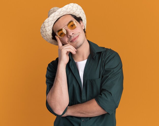 Feliz joven en camisa verde y sombrero de verano con gafas mirando al frente con una sonrisa en la cara de pie sobre la pared naranja