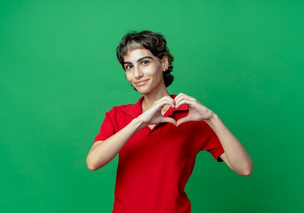 Feliz joven caucásica con corte de pelo pixie haciendo signo de corazón aislado sobre fondo verde con espacio de copia