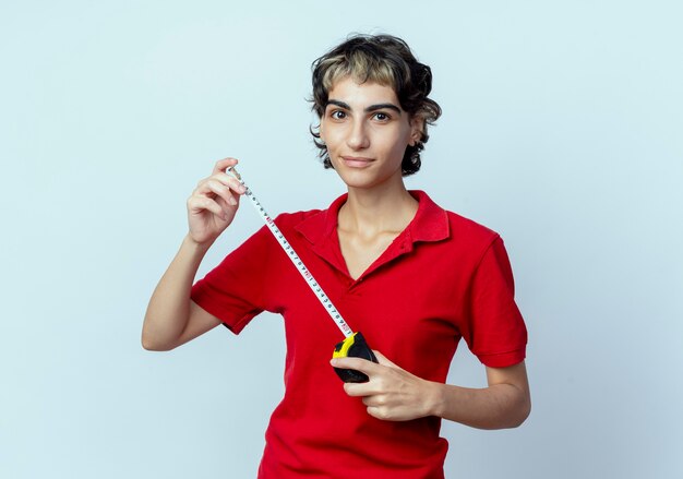 Feliz joven caucásica con corte de pelo pixie sosteniendo medidor de cinta aislado sobre fondo blanco.