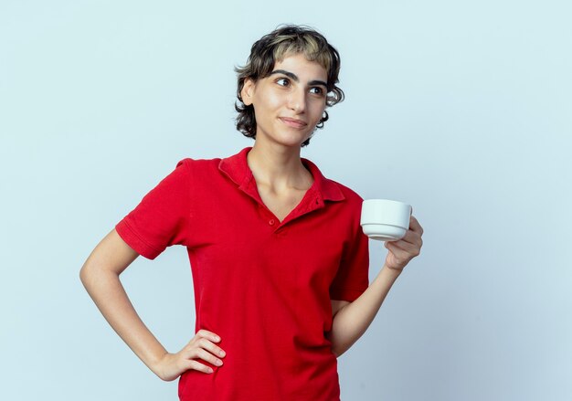 Feliz joven caucásica con corte de pelo pixie sosteniendo la taza mirando al lado aislado sobre fondo blanco con espacio de copia