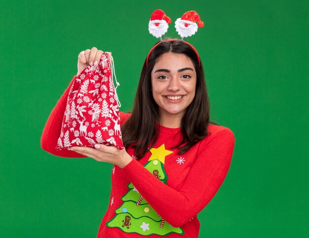 Feliz joven caucásica con diadema de santa sostiene una bolsa de regalo de navidad aislada sobre fondo verde con espacio de copia