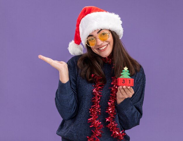 Feliz joven caucásica con gafas de sol con gorro de Papá Noel y guirnalda alrededor del cuello sostiene el adorno del árbol de Navidad y mantiene la mano abierta aislada en la pared púrpura con espacio de copia