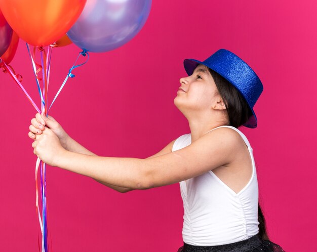 Feliz joven caucásica con gorro de fiesta azul sosteniendo y mirando globos de helio aislados en la pared rosa con espacio de copia