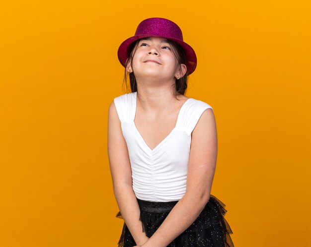 Feliz joven caucásica con gorro de fiesta púrpura mirando hacia arriba aislado en la pared naranja con espacio de copia