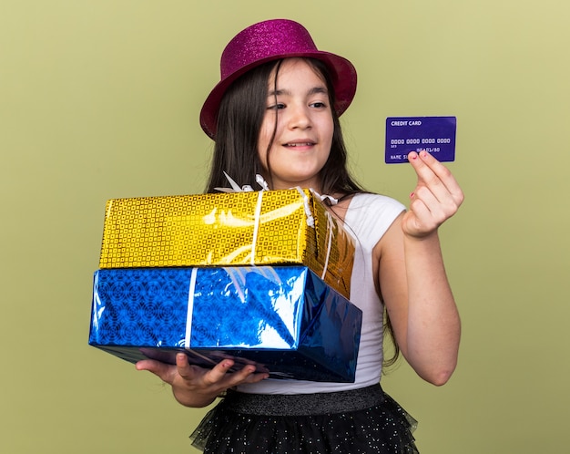 Feliz joven caucásica con gorro de fiesta púrpura sosteniendo cajas de regalo y mirando la tarjeta de crédito aislada en la pared verde oliva con espacio de copia