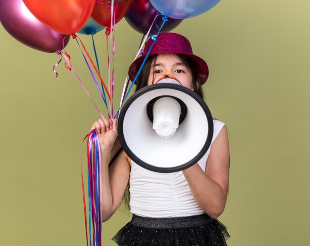 Feliz joven caucásica con gorro de fiesta púrpura sosteniendo globos de helio y altavoz aislado en la pared verde oliva con espacio de copia