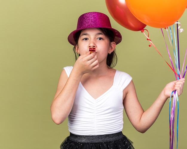 Feliz joven caucásica con gorro de fiesta púrpura sosteniendo globos de helio y soplando silbato de fiesta aislado en la pared verde oliva con espacio de copia