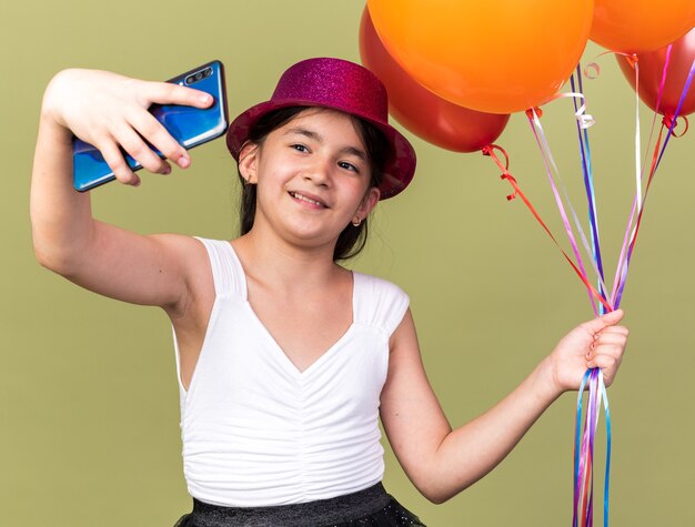 Foto gratuita feliz joven caucásica con gorro de fiesta púrpura sosteniendo globos de helio y tomando selfie en teléfono aislado en la pared verde oliva con espacio de copia