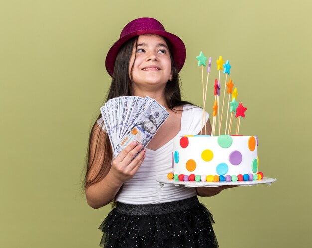 Feliz joven caucásica con gorro de fiesta púrpura sosteniendo pastel de cumpleaños y dinero mirando al lado aislado en la pared verde oliva con espacio de copia