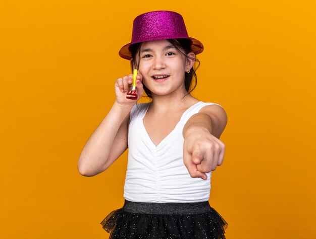 Feliz joven caucásica con gorro de fiesta púrpura sosteniendo el silbato del partido y apuntando aislado en la pared naranja con espacio de copia