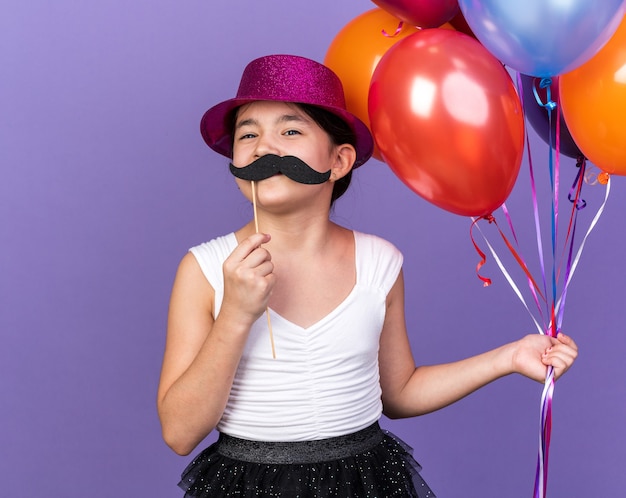 Feliz joven caucásica con gorro de fiesta violeta sosteniendo globos de helio y bigote falso en palo aislado en pared púrpura con espacio de copia